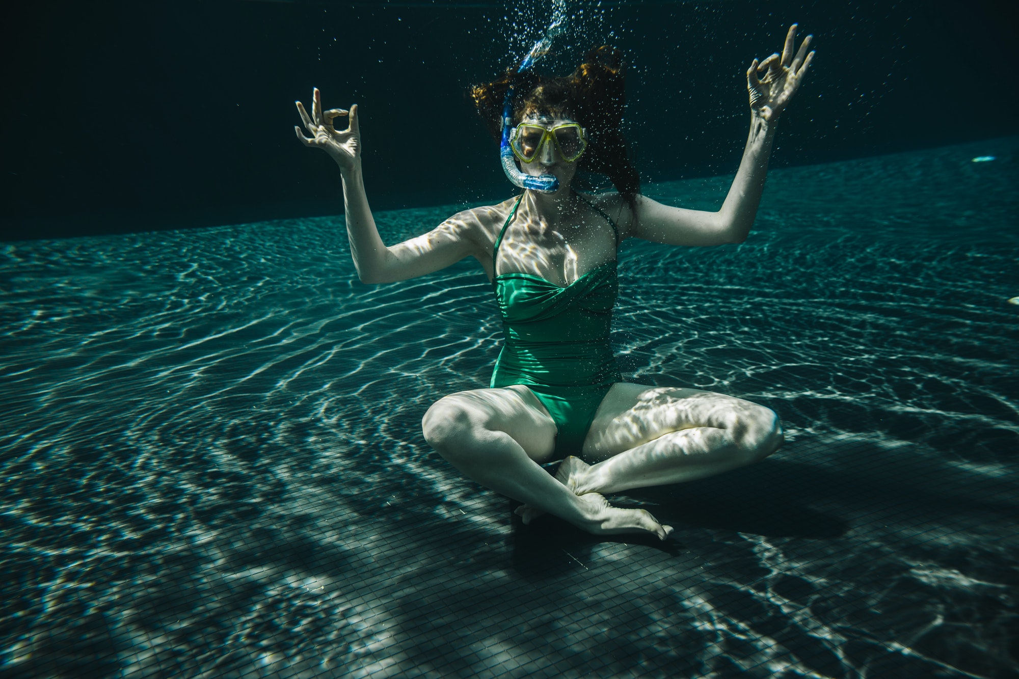 Woman with diving goggles and snorkel sitting in yoga pose underwater in a swimming pool