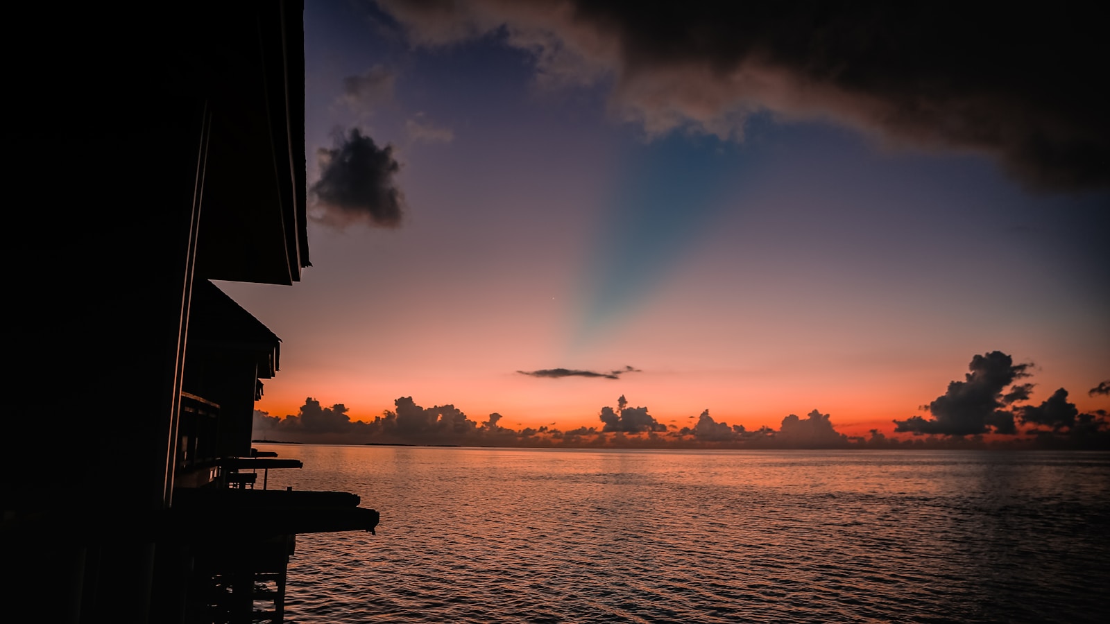 An overwater bungalow with a thatched roof at sunset over calm tropical water